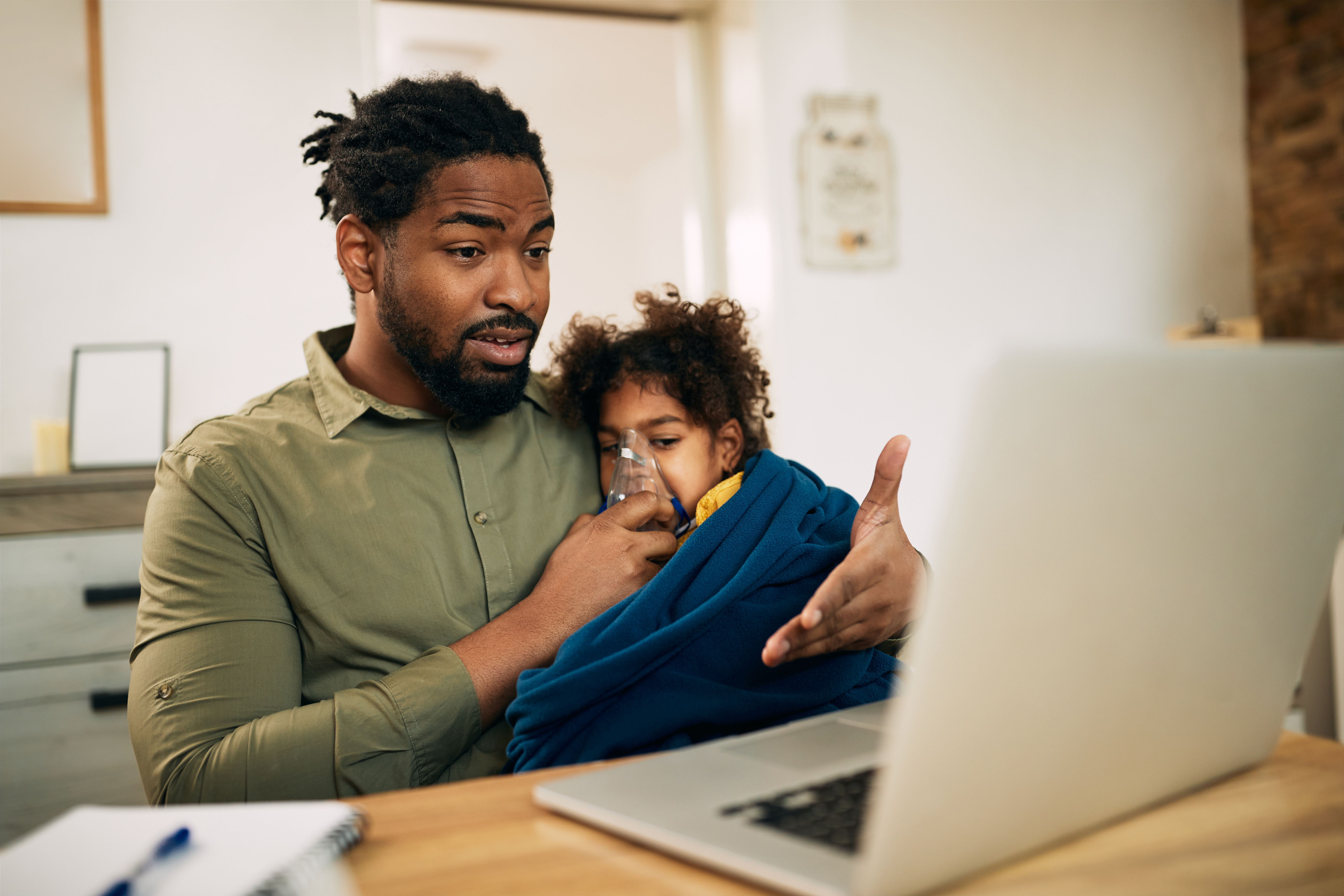 Image of an African American single father giving his daughter inhaling therapy while making video call over laptop at home.